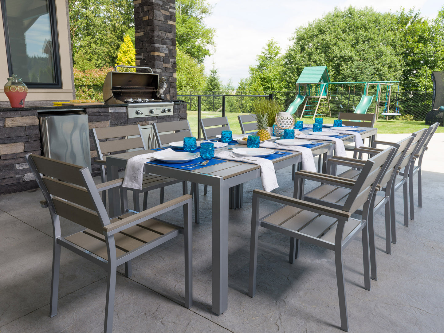 Modern patio dining set with rectangular wooden table, six grey cushioned chairs, and black metal frames, set on a stone patio with greenery in the background. Ideal for outdoor dining and entertaining.