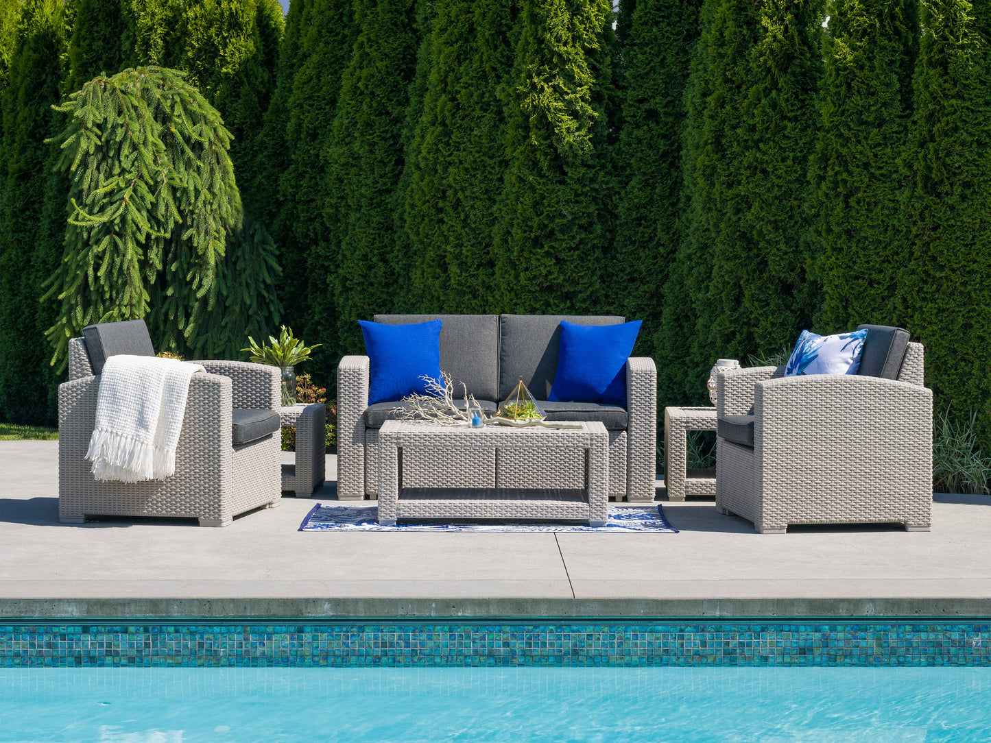 Outdoor patio conversation set with a modern design, featuring a gray wicker loveseat, two matching armchairs, and a glass-top coffee table on a wooden deck, accented with blue and white cushions and throw pillows.