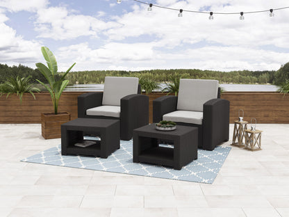 Gray wicker patio conversation set with beige cushions, featuring a loveseat, two armchairs, and a glass-top coffee table, arranged on a wooden deck with greenery in the background.